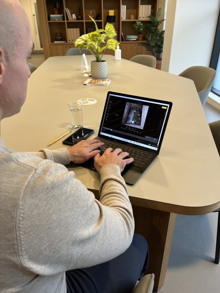 A UK Home Surveyor builds a survey report on ScribeWare UK software on his black laptop. He sits in a gray jumper at a dinning room table, with a glass of water and his cell phone