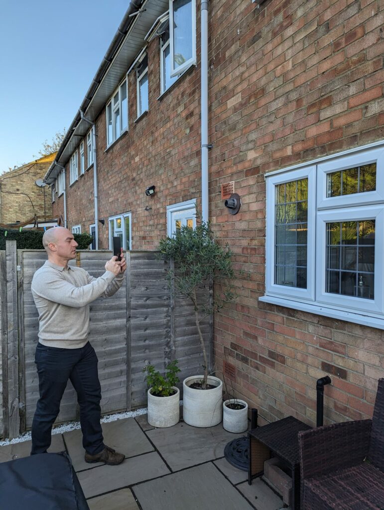 A UK Home Surveyor builds a survey report on ScribeWare UK software by taking photos of a brick house with his black cell phone. He wears black pants and a gray jumper.