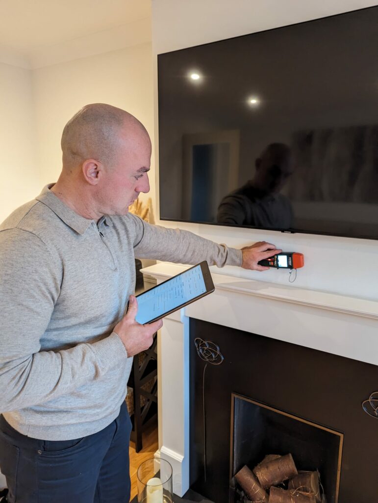 A UK Home Surveyor builds a survey report on ScribeWare UK software using his black tablet. He is taking measurements with a tool held to the wall under a TV.
