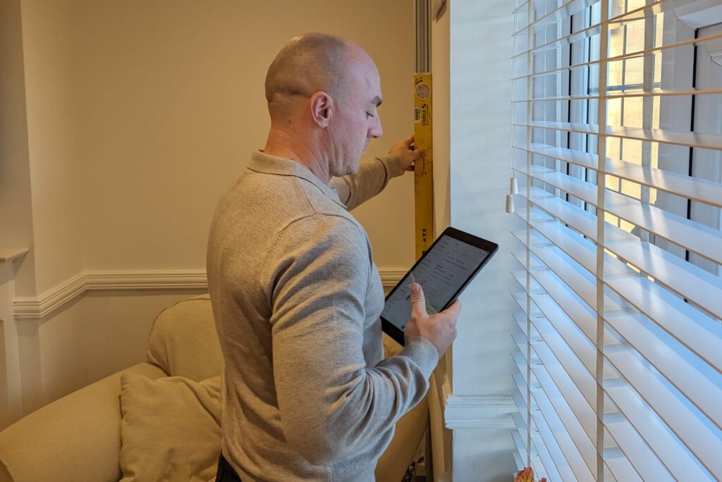 A UK home surveyor in a gray jumper uses a level to check a wall next to a window. He is building a home survey report using ScribeWare UK survey report software on his tablet.