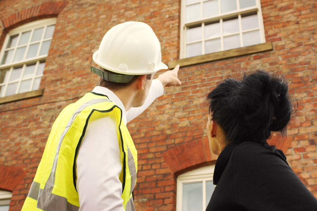 A surveyor points to a windowsill during a survey in the UK. Next to him is a woman with black hair, also looking at the windowsill